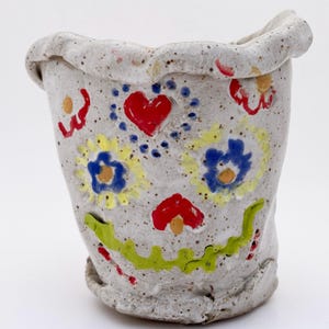 May include: A handmade ceramic cup with a speckled, off-white finish. The cup is decorated with colorful hand-painted designs, including red hearts, yellow and blue flowers, and a green smiling mouth. The rim has a wavy, textured edge.