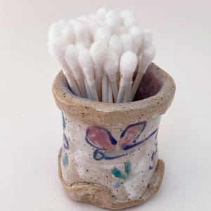 May include: A ceramic holder with a floral design in shades of pink, blue, and green, filled with white cotton swabs.