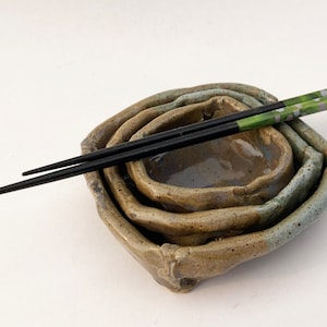 May include: A set of four nesting ceramic bowls in shades of brown and blue-grey, with a pair of black chopsticks resting across the top. The chopsticks have green and purple floral accents on the ends. The bowls have a rustic, handmade appearance.