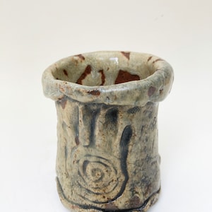 May include: A small, hand-thrown ceramic pot with a rough, textured surface. The pot is glazed in a light green color with brown accents. It has a unique, organic shape and a slightly uneven rim.