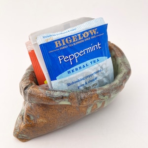 May include: A ceramic tea bag holder shaped like a brown and green sack with a blue and white Biglow Peppermint Herbal Tea tea bag inside. The tea bag reads "Unlock the freshness & enjoy! Biglow. Family Tea Blenders Since 1945. Peppermint. Herbal Tea. Refreshing peppermint... pure & simple. Caffeine Free."