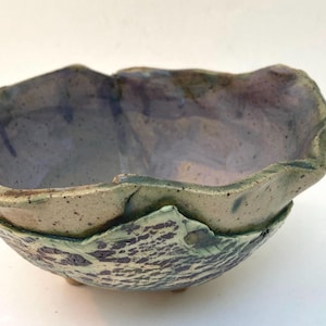 May include: A handmade ceramic bowl with an irregular, wavy rim. The interior features a smooth, glossy lavender glaze with dark speckles. The exterior has a textured, mottled finish in shades of sage green and deep purple, resting on small feet.