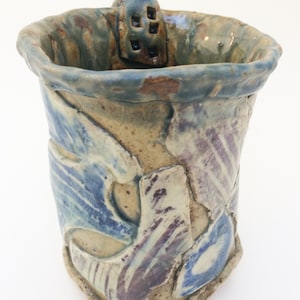 May include: A ceramic mug with a blue, purple, and brown glaze. The mug has a unique, textured design and a small handle with a decorative cutout.