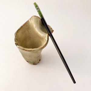 May include: A ceramic chopstick holder with a textured, speckled surface in shades of beige and brown. A black chopstick with a green and white design is resting in the holder.