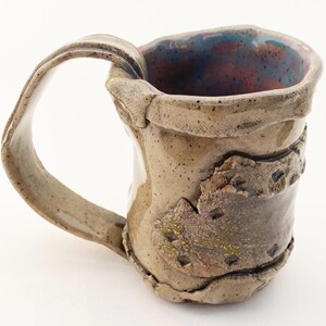 May include: Handmade ceramic mug with a textured, earthy appearance. The mug features a large, curved handle and a unique design with a band of textured clay around the middle. The interior has a blue and red glaze.