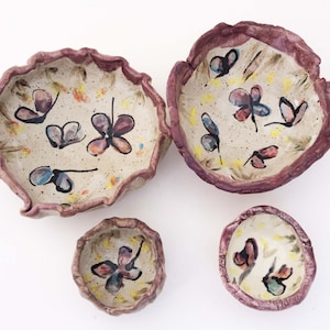 May include: Three ceramic bowls with a white background and a pink rim. The bowls are decorated with hand-painted butterflies in shades of pink, blue, and yellow.