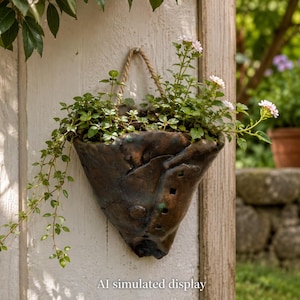 May include: A dark brown, textured ceramic wall planter with a triangular, organic shape and a rope hanger. The planter contains a green leafy plant with small pink flowers. The text "AI simulated display" is printed at the bottom.