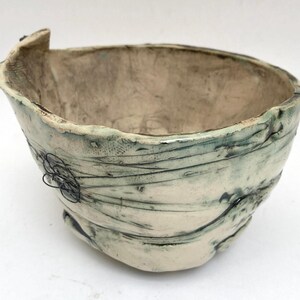 May include: A hand-formed ceramic bowl with an irregular, organic shape and a cream-colored glaze. The exterior features dark green and black linear markings, a textured surface, and a small, decorative black wire coil attached to the side.