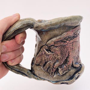 May include: A handmade ceramic mug with a unique, textured, and slightly irregular shape. The mug has a light brown glaze with a pink and brown speckled pattern. The handle is thick and curved.