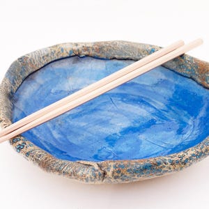 May include: A handcrafted ceramic bowl with a vibrant blue glazed interior and a textured, earthy brown rim. The bowl features an irregular, organic shape and is paired with a pair of light-colored wooden chopsticks resting across the top.