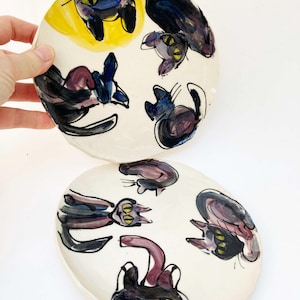 May include: Two white ceramic plates with hand-painted black cat illustrations. The cats have yellow eyes and are depicted in various poses. One plate has a yellow background. The plates are a unique, artistic addition to any home.