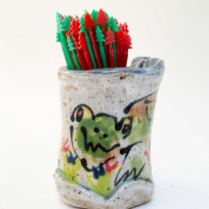 May include: A ceramic toothpick holder with a hand-painted frog design. The holder is filled with red and green tree-shaped toothpicks. The holder has a speckled, off-white finish with colorful accents.