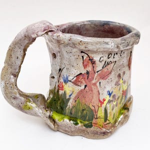 May include: Handmade ceramic mug with a whimsical design. The mug features a textured, speckled exterior with a painted scene of a pink figure in a field of flowers. The handle is thick and organically shaped. The mug has the word "cool" written on it.