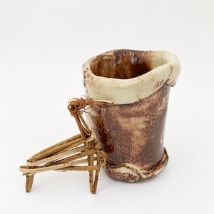 May include: A small, brown and white ceramic pot with a unique, irregular shape. The pot is sitting on a wooden stand with three legs.