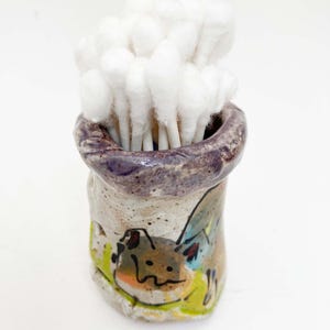 May include: A handmade ceramic container with a cartoon illustration, filled with white cotton swabs. The container has a purple rim and a whimsical design, perfect for bathroom organization. The container is approximately 4 inches tall.