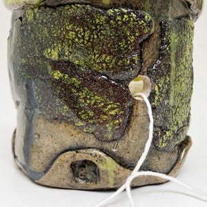 May include: A handmade ceramic pot with a textured surface. The pot features a blend of earthy tones, including brown, green, and yellow. A white string is attached to the side. The pot's design suggests a rustic, artisanal aesthetic.