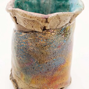 May include: A handmade ceramic vase with a unique, textured surface. The vase features a cylindrical shape with a flared, irregular rim. The exterior showcases a blend of colors, including blue, yellow, and red, with a turquoise interior. The vase is approximately 5 inches tall.