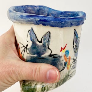 May include: A white ceramic planter with a blue rim and a hand-painted design of two blue cats with colorful flowers and leaves.