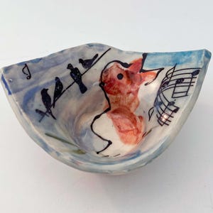 May include: A small, white ceramic bowl with a blue and white glaze. The bowl has a whimsical design featuring a red rabbit singing with birds on a wire and musical notes.