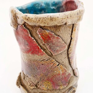 May include: A handmade ceramic vase with a textured surface. The vase features a beige exterior with red, orange, and purple accents. The interior rim is beige, and the inside is blue and red. The vase is approximately 5 inches tall.