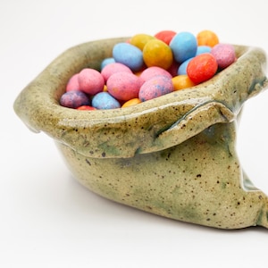 May include: A green ceramic bowl shaped like a bag, filled with colorful candy eggs.