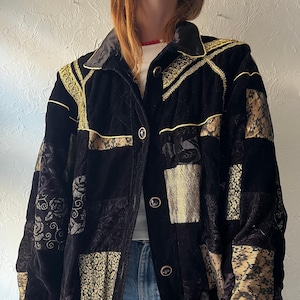 Peut inclure: Une veste noire présentant un motif patchwork composé de panneaux en velours et en dentelle noirs et dorés. Le vêtement comprend un col en satin, une bordure métallique dorée sur les épaules et les poignets, et quatre boutons circulaires.