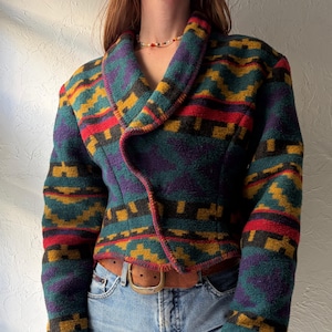 Peut inclure: Veste à motifs colorés avec col châle. Le motif géométrique est composé de teintes de teal, violet, rouge et or. La veste est portée avec un jean bleu clair et une ceinture marron. Le design rappelle une couverture traditionnelle.