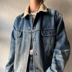 May include: A blue denim jacket featuring a cream-colored sherpa collar, long sleeves, and a front button closure. The jacket includes two chest pockets with button flaps and metallic hardware. The denim fabric has a faded wash finish throughout.