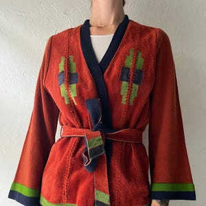 May include: A rust-colored jacket featuring a dark navy blue trim along the V-neckline and hem. The jacket includes a matching tie belt, long sleeves with green and navy cuffs, and geometric green and navy patches on the chest area.