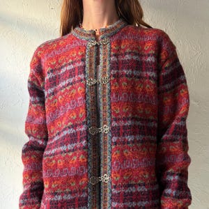 May include: A patterned cardigan in red, orange, blue, and purple. The sweater has a round neckline and decorative metal clasps. The sleeves and hem have a ribbed design. The garment is made of a thick, textured material.