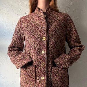 Peut inclure: Une veste matelassée avec un motif cachemire rose foncé, marron et vert olive. La veste présente un col montant, des manches longues, une fermeture boutonnée à l'avant avec des boutons clairs et deux grandes poches plaquées avec bordure marron.