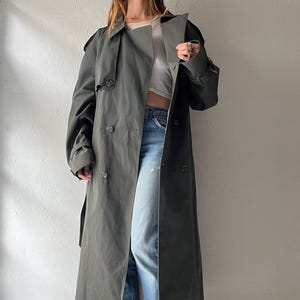 May include: An olive green double-breasted trench coat with a wide collar, shoulder epaulets, and button closures, worn over a white cropped top, light blue denim jeans, and dark pink open-toe heeled mules with wooden platform soles.