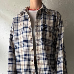 May include: A long-sleeved plaid shirt in navy blue, beige, and brown. The shirt features a button-down front, a collared neckline, and buttoned cuffs. The shirt is worn open over a white t-shirt and blue jeans.