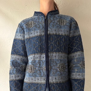 May include: A blue quilted jacket with a stand-up collar and a full-length zip. The jacket features a repeating pattern in shades of blue and grey. The jacket is worn over blue jeans.