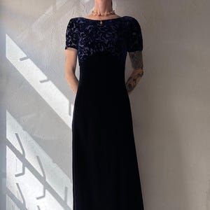 May include: A long, navy blue velvet dress. The bodice has a dark blue floral pattern on the upper section and short sleeves. The dress is worn with striped pointed-toe heels. The dress is suitable for formal occasions.