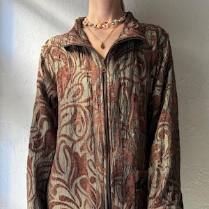 May include: A patterned zip-up jacket in shades of brown and olive green. The jacket features a high collar, a full-length zip, and a textured fabric. It has a floral design.