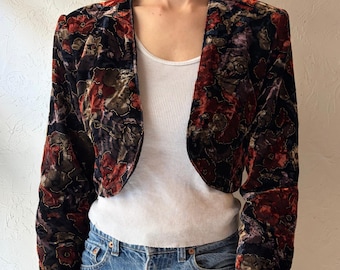Puede incluir: Chaqueta bolero de terciopelo vintage con un estampado floral en tonos rojos, marrones y negros. La chaqueta tiene mangas largas y un diseño corto y abierto. La modelo lleva una camiseta sin mangas blanca y vaqueros azules.
