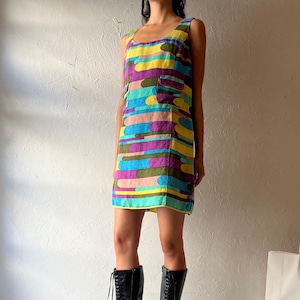 May include: A sleeveless, knee-length dress with a colourful, horizontal, abstract pattern in yellow, blue, purple, and green. The dress is paired with black, lace-up, knee-high boots with stiletto heels. The boots have pointed toes.