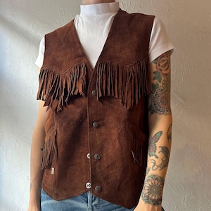 Può includere: Un gilet in pelle scamosciata marrone con frange sul petto e sulle tasche. Il gilet ha una chiusura con bottoni sul davanti e un design a V. Il gilet è in stile vintage.