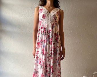 May include: A sleeveless, floor-length floral dress with a white background and red, pink, and blue roses. The dress features a lace-trimmed neckline and a gathered bodice. The model is wearing white socks with lace trim and yellow Mary Jane shoes.