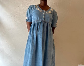 May include: A light blue denim dress with white embroidery around the neckline and on the sleeves. The dress has a gathered waist and a midi length.