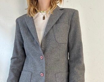 Puede incluir: Una chaqueta gris con dos bolsillos de solapa y tres botones rosas. La chaqueta tiene un corte clásico y una longitud ligeramente recortada.