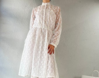 Puede incluir: Un vestido midi de encaje blanco con mangas largas y cuello alto. El vestido tiene un cierre de botones en la parte delantera y una cintura fruncida. La modelo lleva el vestido con botines blancos.