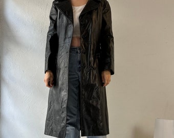 May include: A long black leather coat with a button closure and a collar. The coat is worn over a white crop top and blue jeans.