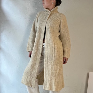 May include: A long, cream-colored knit coat with a high collar and button closure. The coat has a ribbed texture and two front pockets. The person is wearing cream-colored pants and white sneakers.