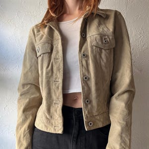May include: A tan suede jacket with two chest pockets and button closures. The jacket has a collared neckline and long sleeves. The jacket is worn with a white top and black jeans.