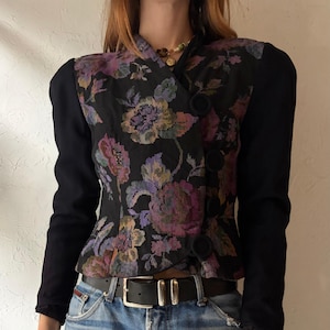 Op de afbeelding: Een zwarte vintage blazer met lange mouwen en een bloemen tapestry design. De blazer heeft een dubbele rij knopen met drie zwarte knopen en een reverskraag. De mouwen zijn zwart en het bloemenpatroon bevat paarse, roze en gele tinten.