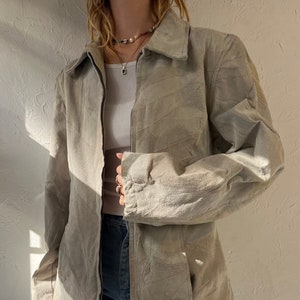 May include: A light gray suede jacket with a zipper closure and a diamond-shaped pattern. The jacket has long sleeves and a collar.
