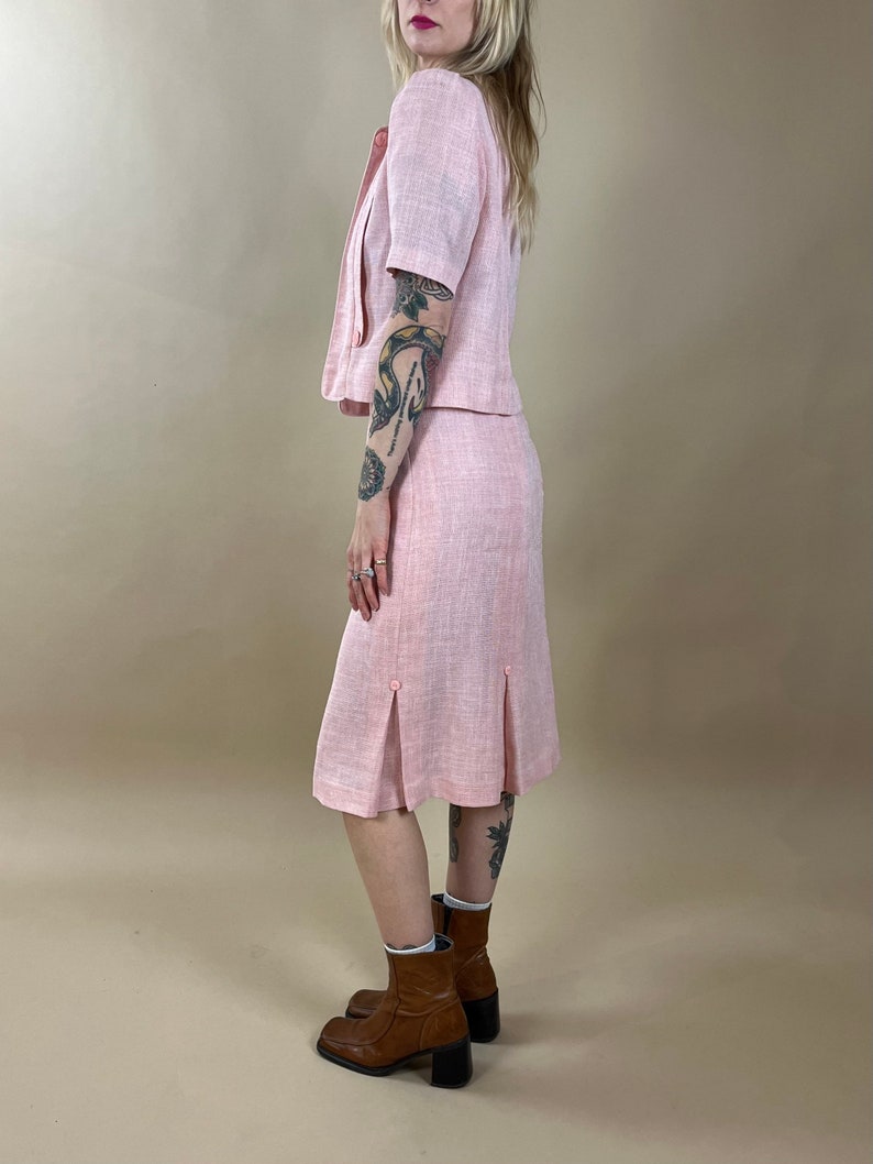 Puede incluir: Un traje de falda de lino rosa con una chaqueta de manga corta y una falda hasta la rodilla. El traje tiene una textura sutil y una silueta cl&aacute;sica y entallada.