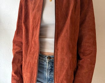 May include: A rust-colored suede jacket with a full-length zipper. The jacket has a collared neckline and long sleeves. The model is wearing a white tank top and blue jeans. The jacket appears to be a vintage or retro style.